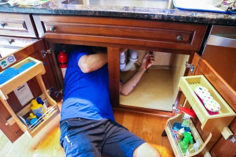 technician in blue shirt repairing sink underneath kitchen cabinet
